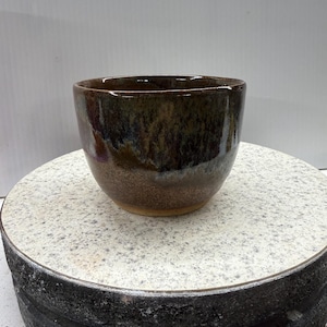 May include: A handcrafted ceramic bowl with a dark brown glaze. The bowl has a rounded shape and a smooth, glossy finish. The interior of the bowl is a lighter brown colour. The bowl is on a speckled white and grey surface.