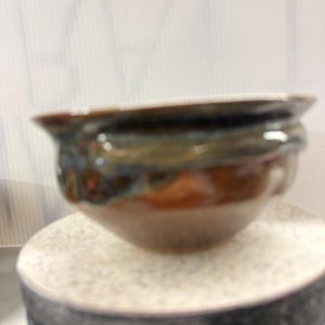 May include: A handcrafted ceramic bowl with a glossy, multi-toned glaze. The bowl features shades of brown, green, and blue, creating a unique and artistic appearance. The rim is slightly flared, and the bowl sits on a speckled surface.
