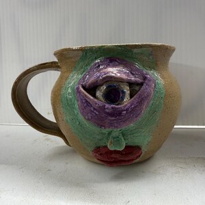 May include: A handmade ceramic mug with a whimsical face design. The mug features a large eye with a purple eyelid, green surrounding area, and red lips. The mug has a tan exterior and a curved handle.