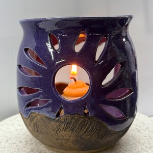 May include: A handcrafted, ceramic candle holder in a deep indigo hue. The holder features leaf-shaped cutouts around the sides, with a textured brown base. An orange candle is lit inside, casting a warm glow.
