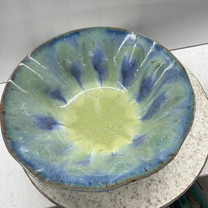 Könnte beinhalten: Eine handgefertigte Keramikschale mit einer einzigartigen, organischen Form. Die Schale weist eine Mischung aus Farben auf, darunter Blau, Grün und Gelb, wodurch ein optisch ansprechendes Design entsteht. Der Rand hat eine leicht wellige Kante, die zu ihrer handwerklichen Ästhetik beiträgt.