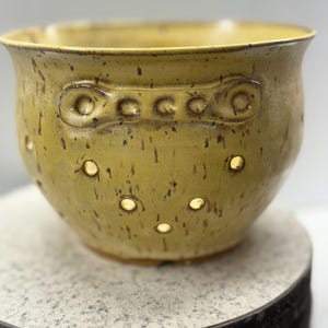 May include: An olive-green ceramic bowl with a textured finish and small, evenly spaced holes. The bowl has a decorative band with circular and oblong shapes. The bowl is placed on a round, speckled surface. The bowl is likely handmade.