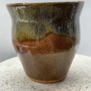May include: A handmade ceramic mug with a distinctive, earthy colour scheme. The mug displays a gradient of colours, including brown, orange, green, and grey. The interior and base are a lighter brown. The mug has a slightly flared rim.