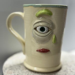 May include: Cream-colored ceramic mug with a sculpted face design. Features a large eye, green eyebrow, green nose, and red lips. The rim has a light green trim, and the handle is curved.