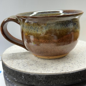 Op de afbeelding: Een handgemaakte keramische mok met een afgeronde vorm en een stevig handvat. De mok heeft een rijk, aards kleurenpalet met tinten bruin, groen en blauw, wat een rustieke en unieke uitstraling creëert. Perfect voor koffie of thee.
