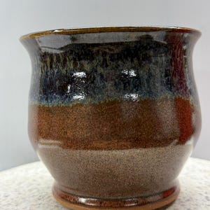 May include: A handcrafted ceramic pot with a glossy glaze. The pot displays a colour gradient, shifting from dark blue at the top to brown and tan at the base. The rim is slightly flared, and the base is rounded.