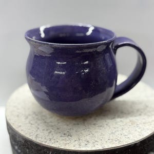 May include: A handcrafted, deep purple ceramic mug with a rounded body and a sturdy handle. The mug has a glossy finish and a slightly textured surface. The mug is sitting on a round, speckled surface.