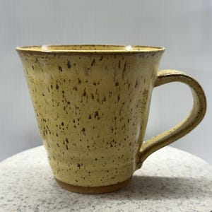 以下が含まれることがあります： 手作りの陶器マグカップ。 黄色の斑点のある釉薬が施されています。 マグカップは幅広で、わずかにフレアしたボディと湾曲したハンドルが付いています。 外側には小さな濃い茶色の斑点があり、素朴な雰囲気を加えています。