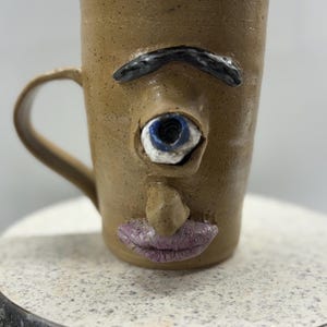 以下が含まれることがあります： 前面に顔が彫刻された、茶色のセラミックマグ。マグには青と白の目、黒い眉、鼻、紫の唇があります。マグには湾曲したハンドルが付いています。