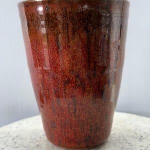 May include: A handmade ceramic tumbler with a rich, reddish-brown glaze. The cup has a slightly tapered shape and a textured surface, showcasing variations in colour from dark brown to a vibrant red. The pottery is suitable for beverages.