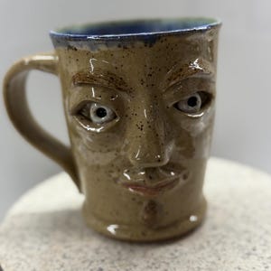 May include: A handmade ceramic mug with a face sculpted on the front. The mug is tan with dark speckles, a blue rim, and a brown handle. The face has white eyes, a nose, and a small mouth.