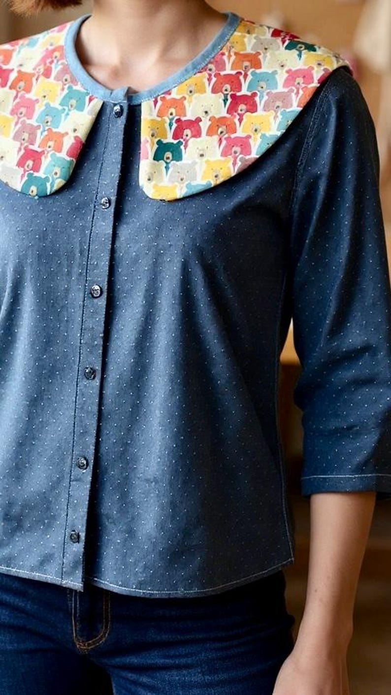 May include: A blue shirt with a white polka dot pattern and a large, rounded collar. The collar is decorated with a colourful bear print. The sleeves are three-quarter length.