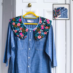 May include: A denim shirt with a detachable floral collar. The collar features a dark background with pink roses, green leaves, and yellow accents. The shirt is button-down with two chest pockets and is hanging on a yellow hanger.