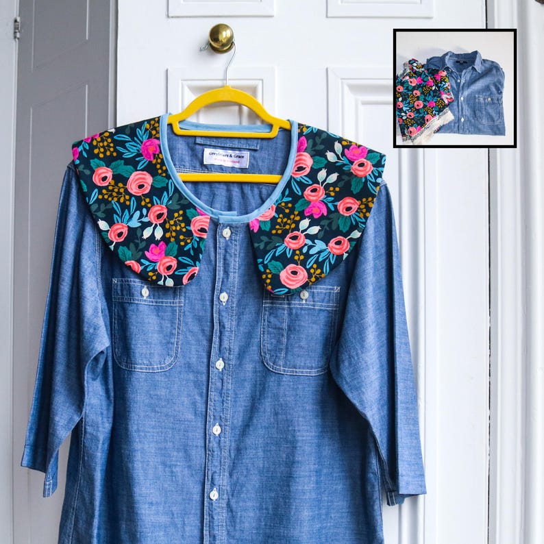 May include: A denim shirt with a detachable floral collar. The collar features a dark background with pink roses, green leaves, and yellow accents. The shirt is button-down with two chest pockets and is hanging on a yellow hanger.