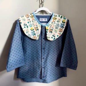 May include: A blue button-down shirt with a floral print collar. The collar features a white background with colorful flowers and green leaves. The shirt has a pattern of small, dark blue shapes.
