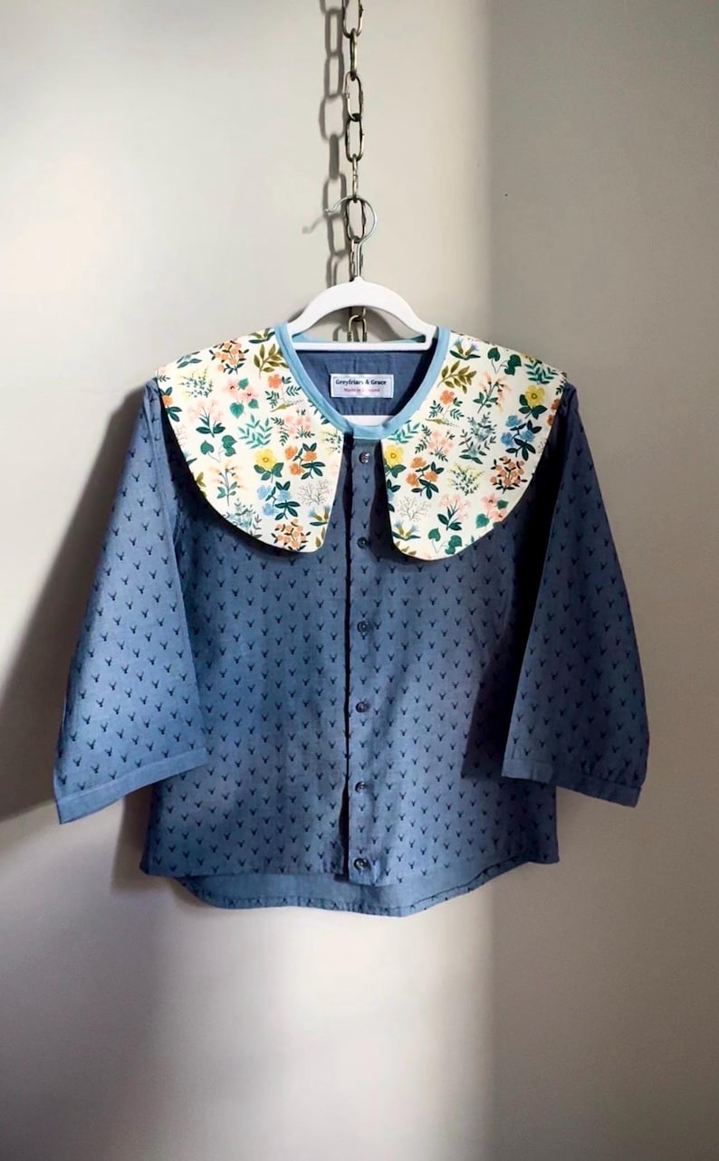 May include: A blue button-down shirt with a floral print collar. The collar features a white background with colorful flowers and green leaves. The shirt has a pattern of small, dark blue shapes.