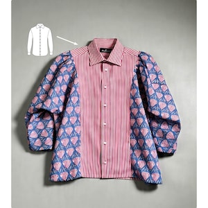 May include: A long-sleeve button-down shirt featuring a central vertical panel with red and white stripes. The sleeves and side panels display a blue fabric with a repeating pink floral pattern. A small white line drawing of a shirt is visible.