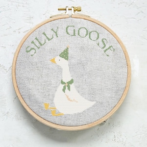 May include: Cross-stitch embroidery in a wooden hoop features a white goose wearing a green party hat and bow tie. The words "SILLY GOOSE" are stitched in green above the goose. The background is a light gray fabric.