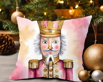 May include: A pink and gold watercolor illustration of a nutcracker soldier on a throw pillow. The nutcracker has a white beard and mustache, a gold crown, and a red and gold uniform.