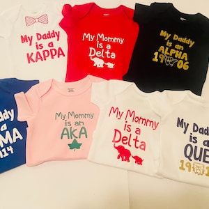 May include: A collection of baby onesies in various colors, including white, red, blue, pink, and black. Each onesie features text such as "My Daddy is a Kappa" or "My Mommy is a Delta" in different fonts and colors. Some designs include graphic elements.