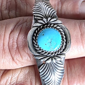 Puede incluir: Una pulsera de plata con una piedra turquesa azul brillante redonda engastada en un diseño de cuerda trenzada. La pulsera tiene diseños de plumas detallados a cada lado.