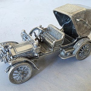 May include: A detailed silver-colored model of a vintage car, featuring a convertible top, spoked wheels, and intricate detailing. The car has a bench seat and a steering wheel. The model is a collectible item.