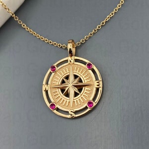 May include: A gold compass necklace with a red gemstone accent at each cardinal direction. The compass features a detailed design with a star in the center.