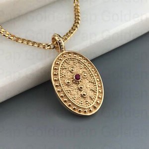 Peut inclure: Un collier en chaîne dorée avec un pendentif ovale présentant un motif texturé et une petite pierre précieuse rouge au centre.