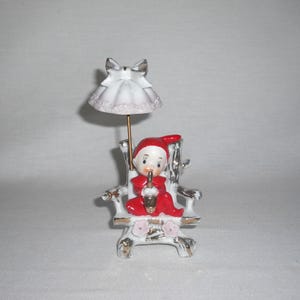 May include: A vintage porcelain figurine of a seated elf wearing a red outfit and hat, holding a gold object. The elf is seated on a white and gold chair with pink floral accents. A white parasol with a bow is above the elf.