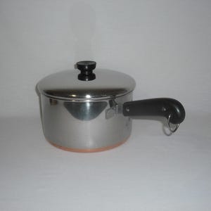 May include: A stainless steel saucepan with a copper-colored base, a matching stainless steel lid with a black knob, and a black handle featuring a small metal hanging loop. The pot has a polished, reflective finish and a cylindrical shape.