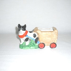 May include: A ceramic figurine of a black and white dog pulling a tan wagon with red wheels. The dog has a red collar and a blue harness. The figurine is on a patch of green grass.