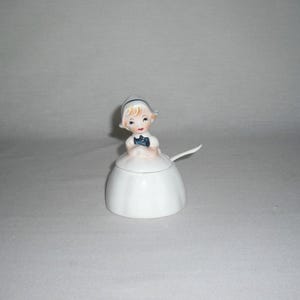 May include: A white ceramic sugar bowl with a lid, shaped like a woman in a dress. The woman has blonde hair, a blue ribbon, and a blue bow. A small spoon rests on the side. A decorative kitchen item.