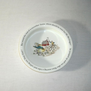 May include: White ceramic plate featuring a scene with a rabbit, a man, and a garden. The plate has text around the rim. The illustration depicts a rabbit running away from a man in a garden setting. The plate is round.