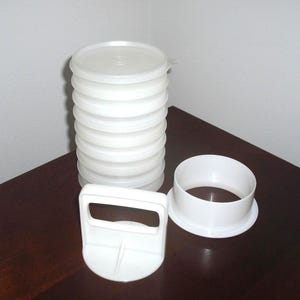 May include: A white plastic kitchen tool set consisting of a tall, cylindrical, stackable container with a lid, a separate circular ring base, and a handheld press component with a rectangular handle, all featuring a smooth, matte finish.