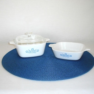 May include: Two white, square casserole dishes with blue floral designs. One dish has a clear glass lid. The dishes are on a blue circular placemat. The casserole dishes have handles.