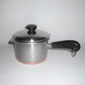 May include: A stainless steel saucepan with a copper-colored base, a black handle featuring a metal hanging loop, and a matching stainless steel lid with a black knob. The pot has a straight-sided cylindrical body and a small pouring spout.