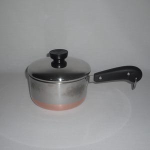 May include: A stainless steel saucepan with a copper-colored base and a matching lid. The pot features a black heat-resistant handle with a small metal loop at the end for hanging and a black knob on the center of the lid for easy lifting.