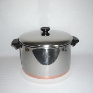 May include: Stainless steel cooking pot with a lid and black handles. The pot has a copper-colored bottom and a black knob on the lid. The pot is designed for cooking and food preparation.