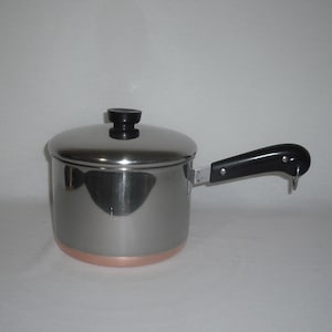 May include: A stainless steel saucepan with a copper-colored base and a matching lid. The pot features a black handle with a small metal loop at the end for hanging. The exterior surface is polished and reflective, showing a cylindrical shape.