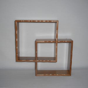 May include: A light brown wooden wall shelf featuring two interconnected square compartments. The structure is composed of smooth, finished wood with a natural grain, creating a geometric display unit with multiple flat surfaces for storage or decor.