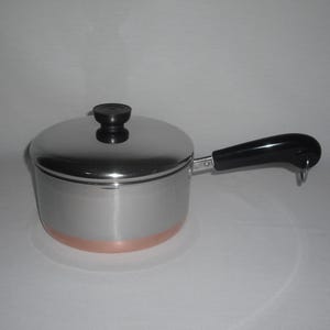 May include: A stainless steel saucepan with a polished finish and a copper-colored base. The pot features a matching stainless steel lid with a black knob and a long, black heat-resistant handle with a small metal loop at the end for hanging.