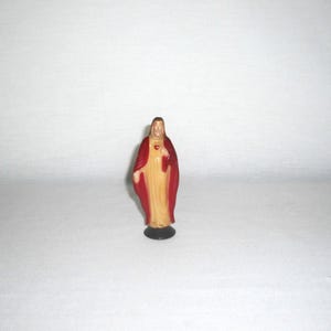 May include: A small standing figurine of a figure with long brown hair, wearing a cream-colored robe and a deep red cloak. The figure has a small red heart emblem on the chest and is mounted on a circular black base, presented against a plain background.