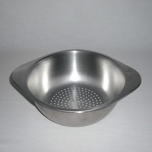 May include: A round, silver-colored stainless steel colander bowl featuring two flat, integrated side handles and a perforated base with numerous small, evenly spaced circular drainage holes for straining liquids from food items.