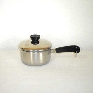 May include: A small, stainless steel saucepan with a lid and a black handle. The lid has a black knob. The handle has a metal hook. The pot is designed for cooking and is suitable for use in a kitchen.