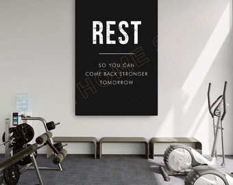 Puede incluir: Póster motivacional en blanco y negro con el texto "REST SO YOU CAN COME BACK STRONGER TOMORROW". El póster está colgado en una pared blanca en un entorno de gimnasio.