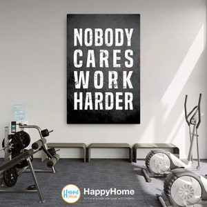 May include: A black and white motivational poster with the text "NOBODY CARES WORK HARDER" in a distressed font. The poster is hanging on a white wall in a gym setting.