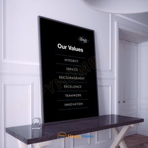 May include: A framed black sign with the text "Our Values" and a list of values in white. The sign is on a dark gray table with a clear glass cube. The background is a white wall and a white door.