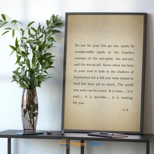 Op de afbeelding: Een ingelijste print met inspirerende tekst in een zwarte lijst. De tekst staat op een beige achtergrond. Links staat een glazen vaas met groene takken op een zwarte tafel.
