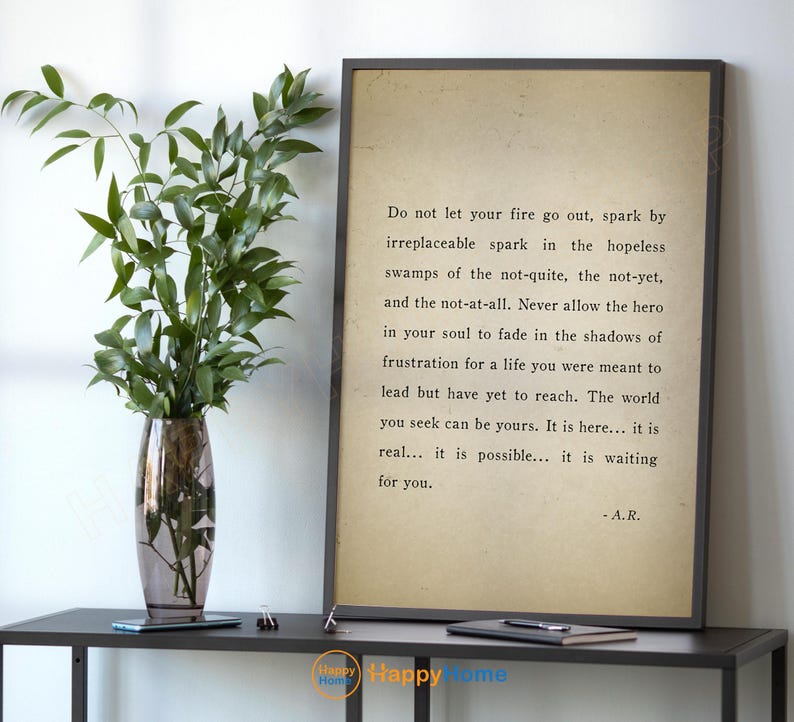 Op de afbeelding: Een ingelijste print met inspirerende tekst in een zwarte lijst. De tekst staat op een beige achtergrond. Links staat een glazen vaas met groene takken op een zwarte tafel.