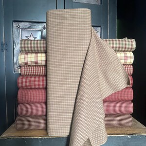 May include: A stack of fabric rolls in various plaid patterns, including red, cream, and brown. A large roll of tan fabric with a grid pattern is in the foreground. The fabrics are stacked on a wooden surface, suggesting a home decor or craft theme.